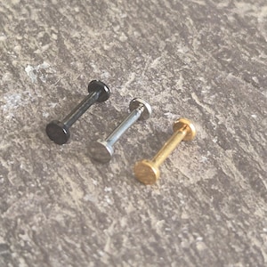 May include: Three flat-back labret studs for body piercings, arranged in a row. The studs feature a straight post with a circular flat disc at each end. One stud is matte black, one is polished silver-tone metal, and one is a gold-tone finish.