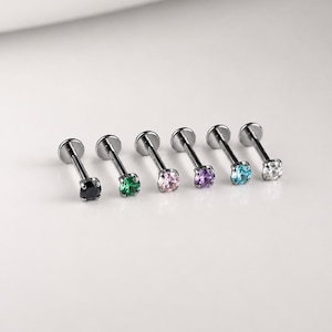 May include: A set of six silver-toned labret studs, each featuring a round prong-set gemstone. The stones are coloured black, green, light pink, purple, light blue, and clear, all attached to a flat circular base with a straight metal post.