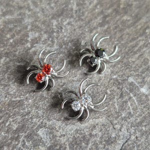 May include: Three silver spider-shaped jewelry pieces with gemstone accents. One spider has red gemstones, another has black, and the third has clear gemstones. The jewelry is displayed on a gray, textured surface.