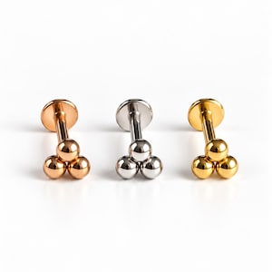 May include: Three labret body jewellery pieces featuring a flat back disc and a front cluster of three small spheres. The set includes one rose gold-tone, one silver-tone, and one gold-tone metal stud, each with a smooth, polished, and reflective finish.