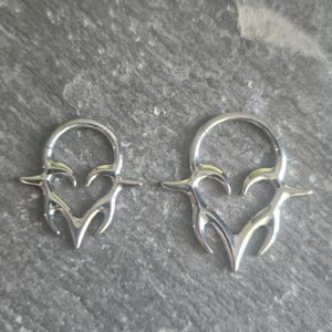May include: Two silver-toned metal hoop rings featuring a decorative heart-shaped design with pointed, flame-like extensions. The jewelry pieces are circular with a hinged closure mechanism, displaying a polished, reflective metallic surface finish.