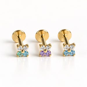 May include: Three gold-tone flat back stud earrings, each featuring a four-stone cluster arranged in a flower shape. The stones include clear, light blue, green, and purple round-cut gems set in a gold-coloured metal frame with a post backing.