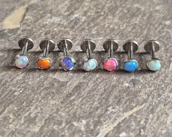 May include: A collection of seven labret studs with colorful opal cabochons. Each stud features a flat, silver-colored backing and post, with the opal stones in shades of pink, orange, blue, and white. The jewelry is displayed on a textured, gray surface.