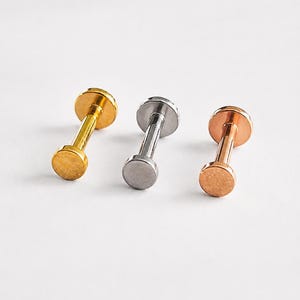 May include: Three flat-back labret studs in gold, silver, and rose gold tones. Each piece features a smooth, circular front disc attached to a straight post and a flat circular base, designed for body piercing applications with a metallic finish.