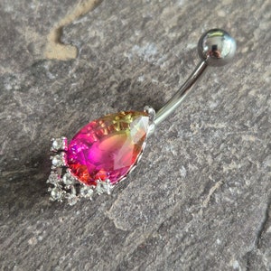 May include: A silver belly button ring with a teardrop-shaped pink and yellow gem surrounded by clear rhinestones.