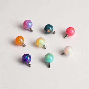 Puede incluir: Ocho accesorios esféricos para joyería corporal con piedras iridiscentes tipo ópalo en varios colores, incluyendo rosa, azul oscuro, naranja, amarillo, morado, azul claro y blanco, cada uno con un pequeño poste metálico roscado.