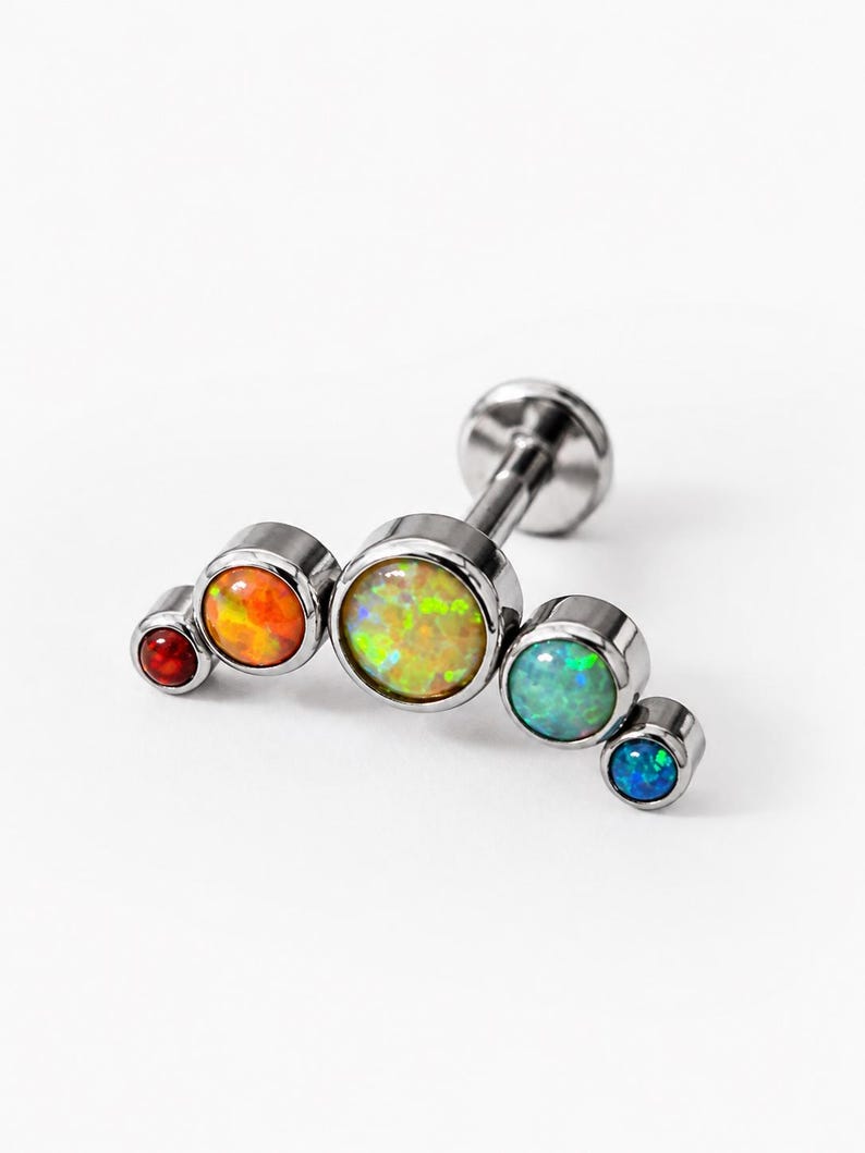 May include: A silver-toned metal body jewellery stud featuring a curved arc design. The piece is set with five round, multi-coloured stones in a gradient of red, orange, yellow, green, and blue, arranged from smallest to largest and back to smallest.