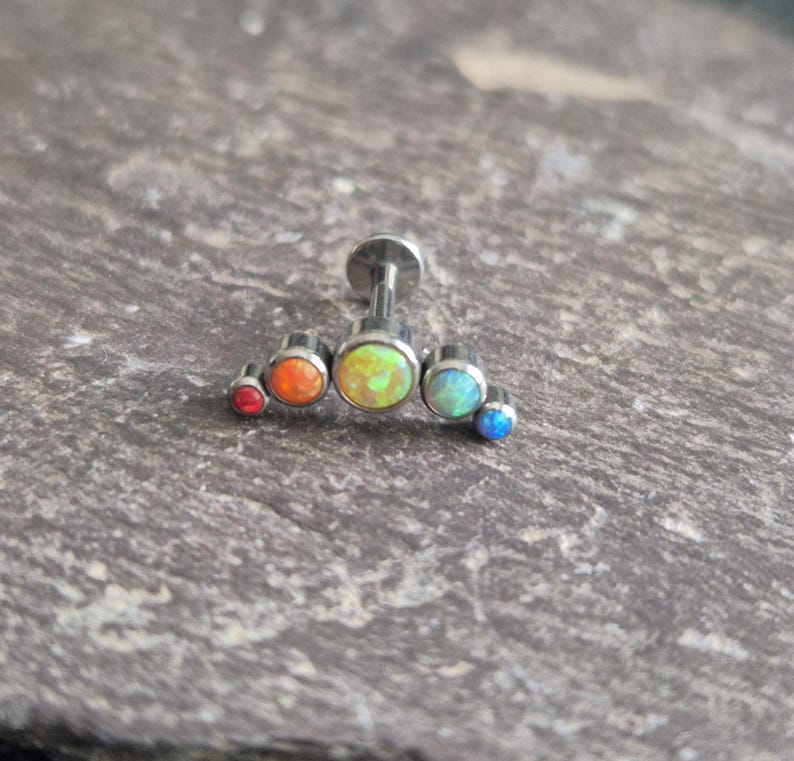 May include: A silver-toned metal body jewellery piece featuring a curved bar design with five round, multi-coloured cabochon stones. The stones transition in colour from a small red gem on the left to orange, a larger central yellow-green, light blue, and a small dark blue gem on the right.