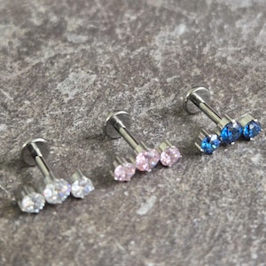 Puede incluir: Tres piercings labiales con parte trasera plana y poste. Un piercing tiene tres cristales transparentes, otro tiene tres cristales rosas y el tercero tiene tres cristales azules. Los piercings son de metal plateado.