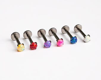 May include: Six labret studs with metallic silver-toned posts and flat back discs. Each stud features a round, prong-set synthetic opal stone in a different color, including yellow, red, purple, pink, blue, and white, arranged in a row.