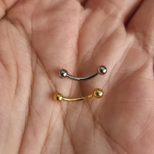 May include: Two curved barbell piercings. One is silver-colored, and the other is gold-colored. Each barbell has a small ball on each end. The jewelry is shown against a skin-toned background.