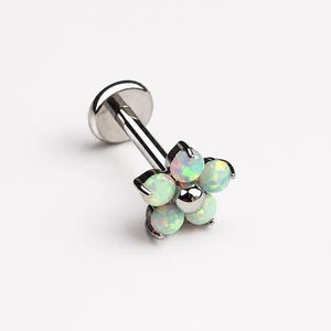 Peut inclure: Un bijou de piercing labret argenté avec une tête en forme de fleur. La fleur se compose de cinq pierres rondes blanc opalin irisé disposées autour d'une perle centrale argentée, fixée sur une tige droite avec un disque plat à l'arrière.