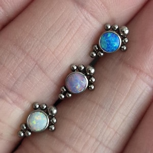 May include: A silver body jewellery piece with three opal stones in shades of blue, lavender, and white. Each stone is surrounded by small silver beads. The piece is on a black bar.