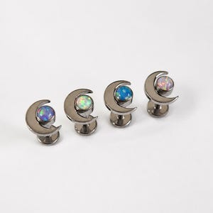 May include: Four dark metal stud earrings, each featuring a crescent moon shape framing a round, iridescent opal cabochon. The opals display varied colour shifts including blue, white, and pink tones, set within a polished, dark-toned metal base.