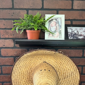 Op de afbeelding: Een bruine geweven zonnehoed met een brede rand zit op een plank voor een bakstenen muur. Een pot met varen en een ingelijste foto van een baby staan ook op de plank.
