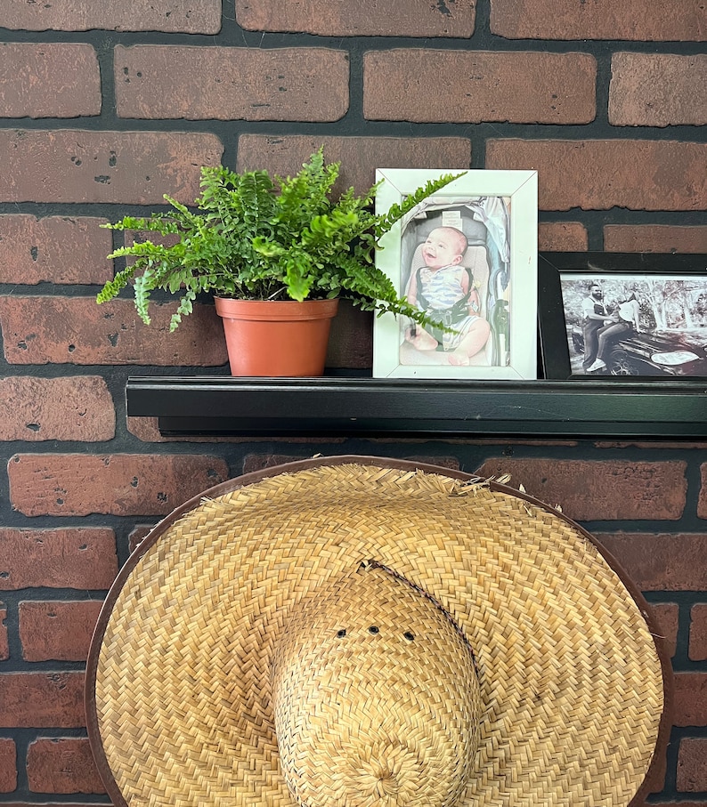 Op de afbeelding: Een bruine geweven zonnehoed met een brede rand zit op een plank voor een bakstenen muur. Een pot met varen en een ingelijste foto van een baby staan ook op de plank.