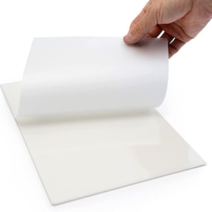 May include: A white sheet of adhesive-backed paper is being peeled back to reveal the sticky side. The paper is smooth and has a slight sheen.
