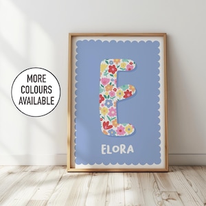 Op de afbeelding: Een ingelijste print met de letter 'E' in een bloemmotief, tegen een lichtblauwe achtergrond met een geschulpte rand. Het bloemmotief bevat rode, gele en roze bloemen. De naam 'ELORA' staat eronder gedrukt.