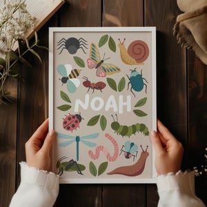 Puede incluir: Un cuadro enmarcado con ilustraciones coloridas de insectos y el nombre "NOAH". Las ilustraciones incluyen una araña, una mariposa, una abeja, una hormiga, una mariquita, una libélula, una oruga, un gusano, un escarabajo, un saltamontes y un caracol.