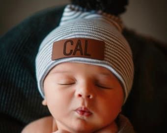 May include: A baby wearing a blue and white striped beanie with a brown leather patch that says "CAL" on it. The beanie has a black pom pom on top.