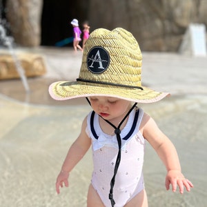 May include: A young child wearing a white swimsuit with a navy blue trim and anchors printed on it. The child is wearing a straw cowboy hat with a pink band and a black patch with the letter "A" on it. The child is standing in shallow water.