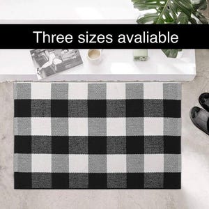 May include: A black, white, and gray checkered doormat. The mat has a large, bold check pattern. The text "Three sizes available" is displayed in a black banner above the mat. The mat is rectangular and appears to be made of woven material.