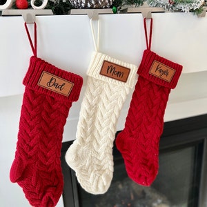 May include: Three red and white knitted Christmas stockings with leather name tags. The stockings are hung on a white mantel with a fireplace in the background. The name tags read "Dad", "Mom", and "Kayla".
