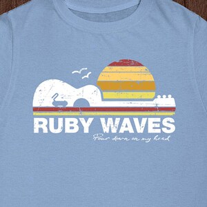 May include: Light blue t-shirt with a vintage-style graphic of a sunset over a guitar with the text "RUBY WAVES" and the words "Pour some on my head" in a handwritten font.
