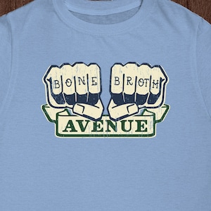 May include: Light blue t-shirt with a vintage-style graphic of two fists with the words "BONE BROTH AVENUE" printed on them.