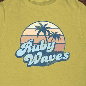 May include: A yellow t-shirt with a retro-style graphic of palm trees and a sunset with the text "Ruby Waves" in blue.