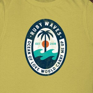 May include: A yellow t-shirt with a distressed, circular logo featuring a palm tree, waves, and the text "Ruby Waves. Ocean of Love Would Carry Me Up. Est. 2008."