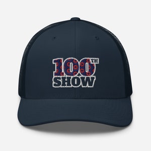 May include: A navy blue trucker hat with a white mesh back. The front of the hat features an embroidered design of the numbers "100" with red and blue donut patterns and the text "TH SHOW" in white.
