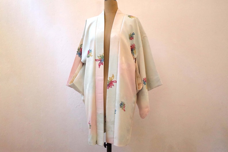 K&ouml;nnte beinhalten: Ein wei&szlig;er und rosa japanischer Kimono mit Blumenstickerei. Der Kimono hat eine hellblaue Unterlage und zeigt bunte Blumen an den &Auml;rmeln und vorne.