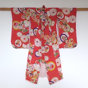 May include: A red kimono with a floral pattern, hanging from a wooden rod. The kimono features a vibrant design with white, yellow, green, and purple circular motifs. The garment's sleeves and body are fully displayed against a white background.