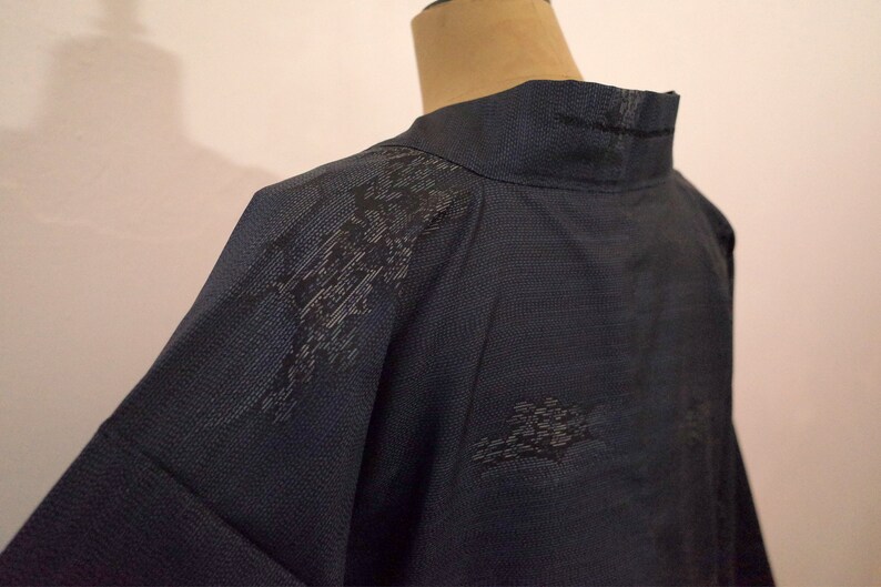 Op de afbeelding: Een donkerblauwe kimono met een zwart bloemenpatroon. De kimono heeft een hoge kraag en lange mouwen.