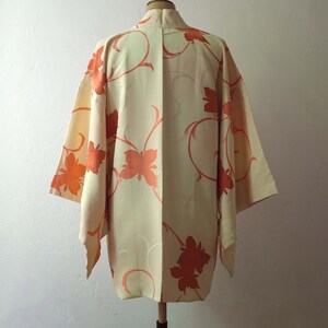 May include: A cream-colored kimono with a floral pattern in shades of orange. The kimono has a traditional Japanese design with a wide collar and long sleeves.