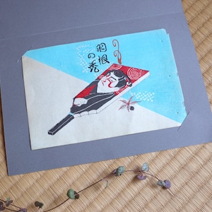 May include: A Japanese-style art print featuring a fan with a stylized face and text. The fan is red, black, and white, set against a blue and white background. The print is inside a gray card.