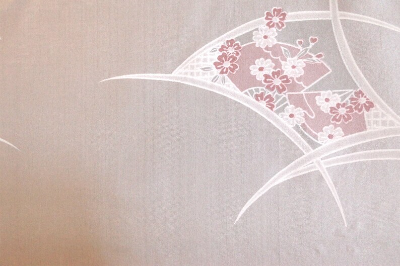 Op de afbeelding: Een witte stof met een delicaat roze bloemenpatroon. Het patroon heeft een waaiervormig ontwerp met witte lijnen en roze bloemen.