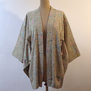 May include: A beige kimono with a floral pattern in shades of pink, blue, and green. The kimono has a wide, open collar and long sleeves.