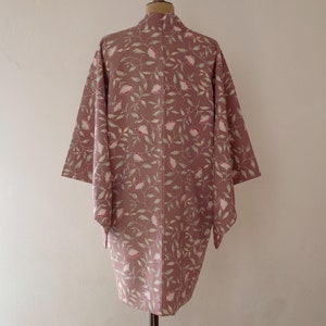 May include: A long, pink kimono with a floral pattern of white flowers and green leaves. The kimono is hanging on a mannequin.