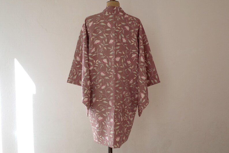 May include: A long, pink kimono with a floral pattern of white flowers and green leaves. The kimono is hanging on a mannequin.