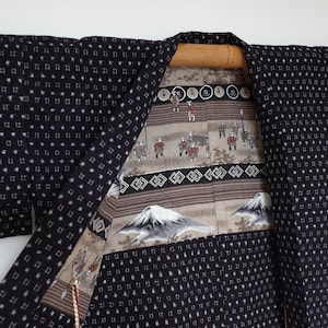 May include: A black and white patterned Japanese Hanten jacket with a brown and white lining. The lining features a mountain scene and a repeating geometric pattern.