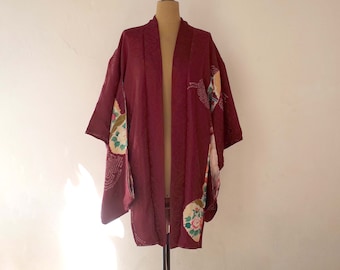Puede incluir: Un kimono burdeos con patrones florales y geométricos. El kimono tiene un cuello ancho y abierto y mangas largas.