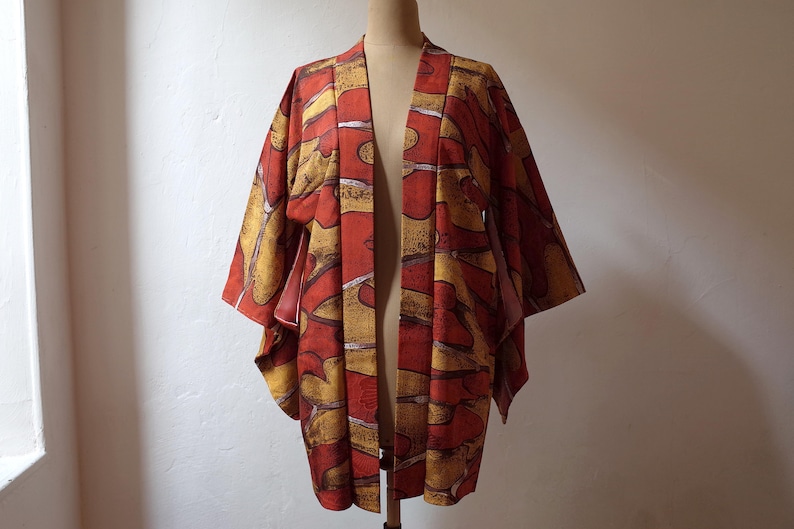 May include: A red and gold Japanese kimono with a geometric pattern. The kimono is open in the front and is hanging on a mannequin.