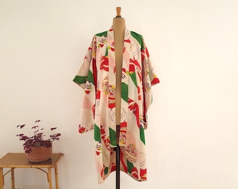 Puede incluir: Una bata kimono vintage en crema, rojo, verde y rosa. La bata se exhibe en un maniquí de madera. El diseño presenta patrones florales y geométricos. Una pequeña mesa de madera con una planta en maceta está en primer plano.