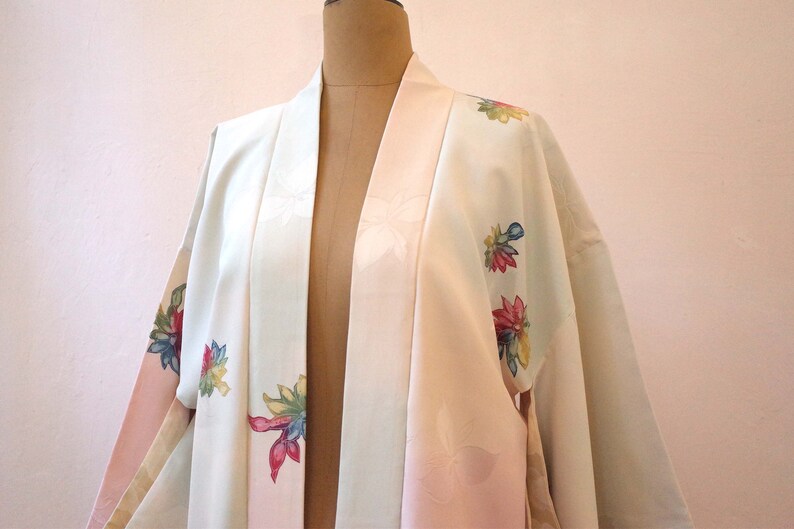 K&ouml;nnte beinhalten: Ein wei&szlig;er und rosa japanischer Kimono mit Blumenstickerei. Der Kimono hat einen rosa G&uuml;rtel und ist mit bunten Blumen verziert.