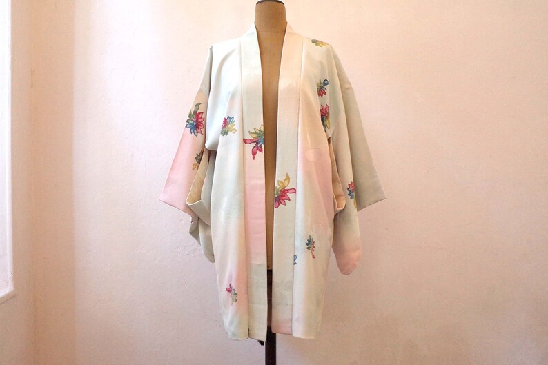 K&ouml;nnte beinhalten: Ein hellblauer und rosa japanischer Kimono mit Blumenmuster. Der Kimono hat einen weiten, offenen Kragen und lange &Auml;rmel. Der Stoff ist weich und seidig.