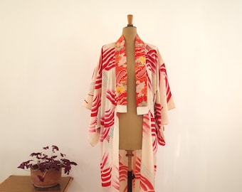 May include: A vintage kimono in cream with red and orange abstract patterns, displayed on a mannequin. The kimono features a floral design on the collar and sleeves. A small potted plant sits on a wooden table in the foreground.