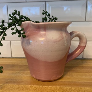 May include: A pink and white ceramic pitcher with a swirl design. The pitcher has a handle and a wide mouth.
