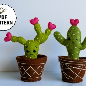 Può includere: Due cactus di peluche in feltro con facce sorridenti e cime a forma di cuore. I cactus sono in vasi marroni con un motivo a rombi. I cactus sono verdi con accenti bianchi e cuori rosa.  MODELLO PDF.